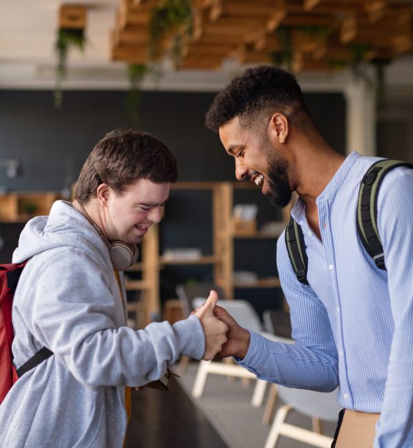 A young happy man with Down syndrome with his mentoring friend greeting indoors at school.