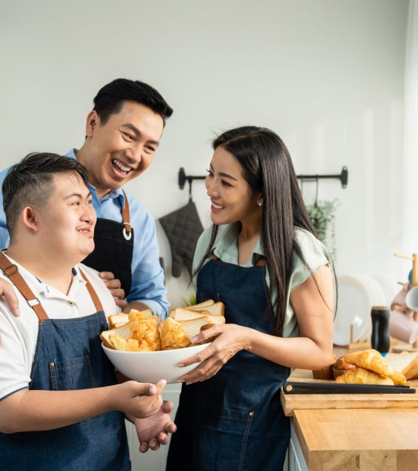 Asian happy family baking bakery with down syndrome's son in kitchen. Beautiful loving mother and father take care and teaching young man with autism or special needs cooking foods indoors at home.